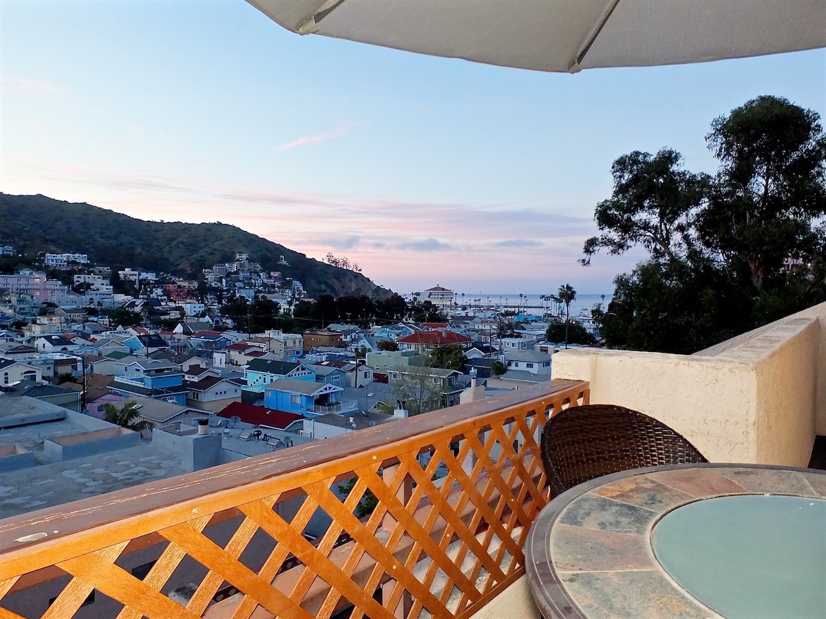 A balcony view showcases a panoramic vista of Avalon, with colorful rooftops and the harbor visible in the distance. A round table with wicker chairs occupies the space, under a large umbrella, while gentle evening hues fill the sky.