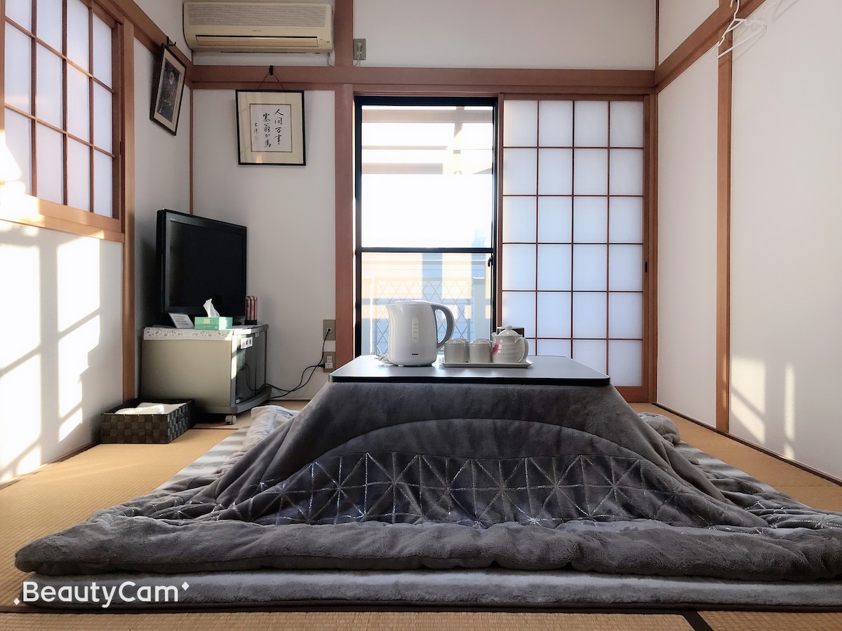 A spacious room is presented with traditional tatami flooring and shoji screens allowing natural light to filter in. A low table is set with a teapot and cups, while a cozy futon lies underneath, creating a comfortable atmosphere for relaxation.