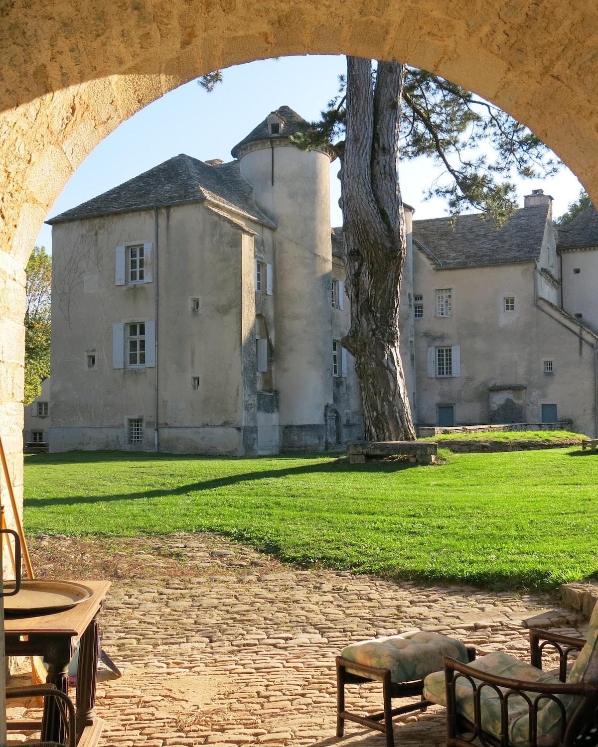 The view from a stone archway overlooks a verdant lawn leading to a historic building, characterized by its unique architectural features. A large tree and cobblestone path frame the scene, providing a tranquil setting in a serene outdoor space.