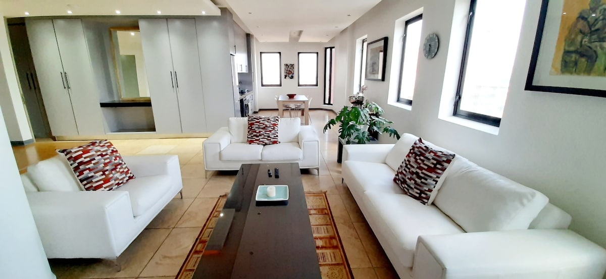 A spacious living area is furnished with modern white sofas adorned with decorative cushions. A coffee table sits centrally on a textured rug, while large windows allow natural light to illuminate the room. The open-plan layout provides a view of the kitchenette in the background.