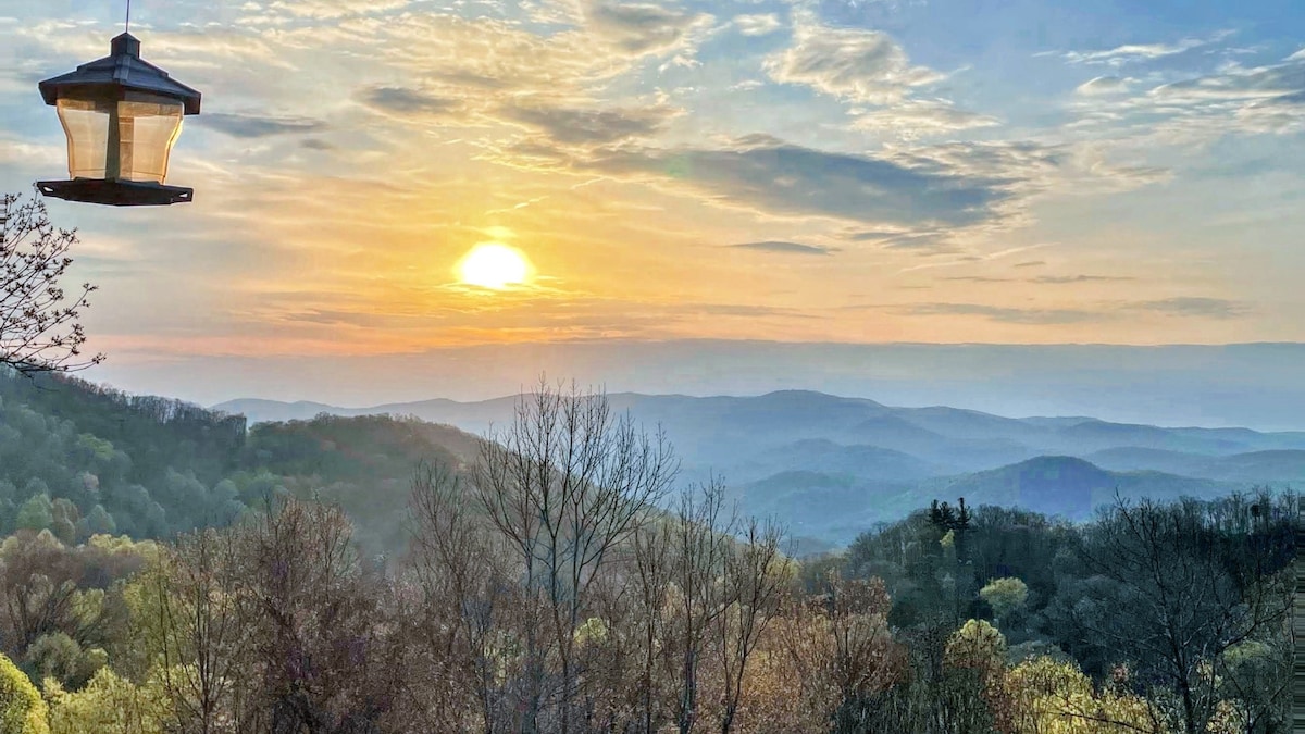 A panoramic view of the mountains during sunrise features soft hues of orange and gold against the horizon. The landscape is layered with rolling hills and a mix of trees, creating a tranquil, natural setting.