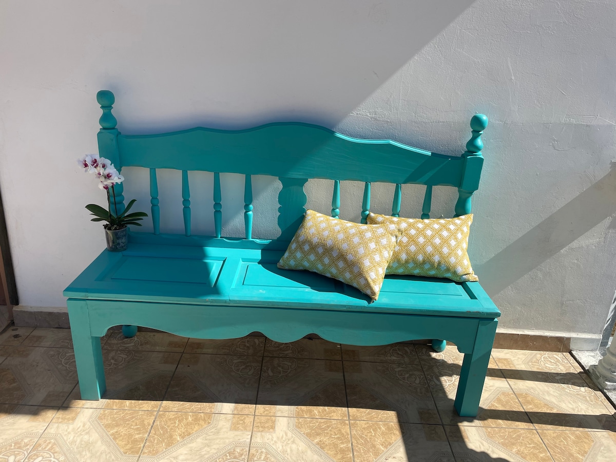 A vibrant turquoise bench is positioned against a white wall, enhanced by the warm sunlight. Two decorative pillows with a yellow and white pattern are placed on the bench, along with a small vase containing an orchid, adding a touch of natural beauty.