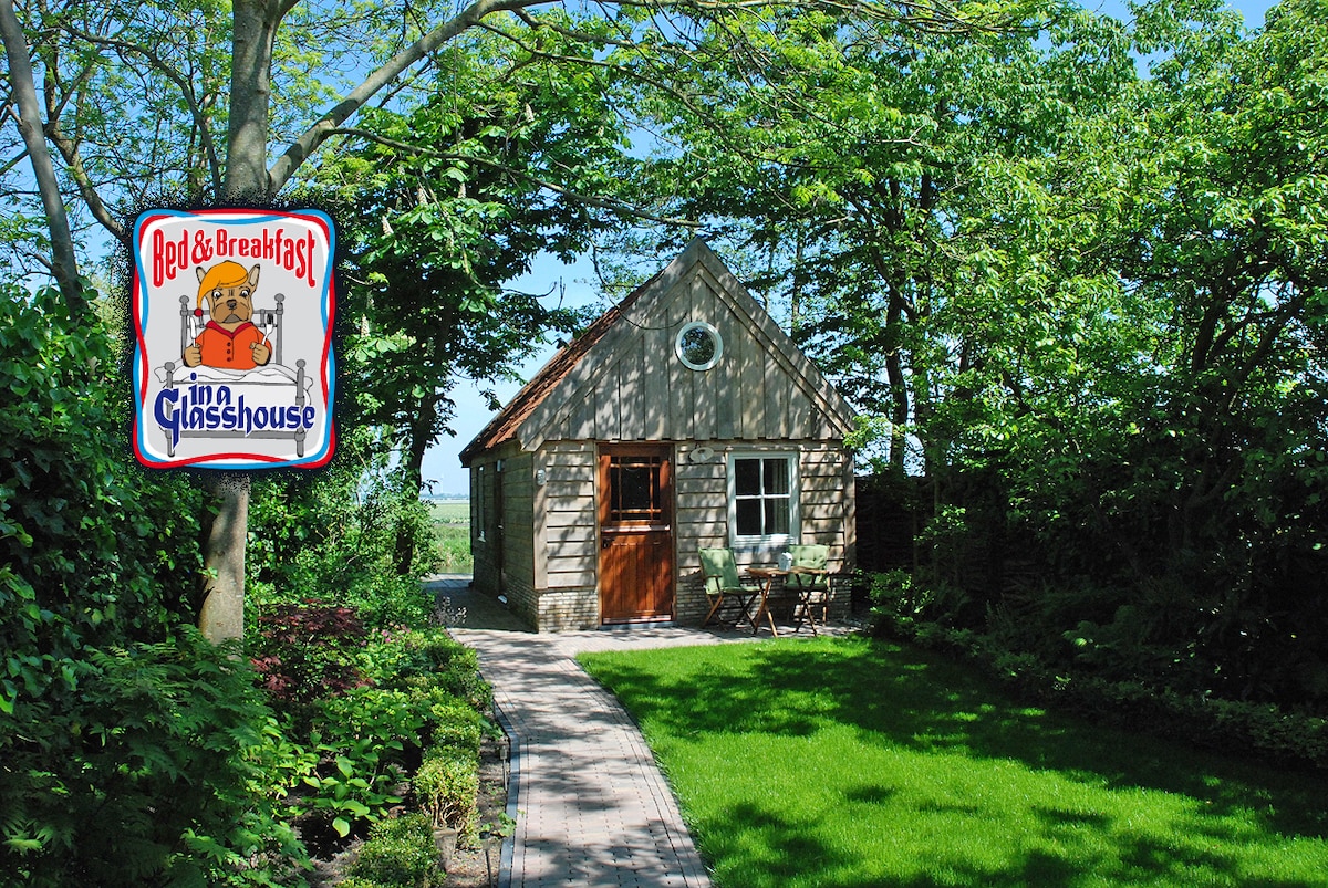 A quaint cottage is set within a lush garden, surrounded by flourishing greenery and trees. A stone pathway leads to the entrance, which features a charming round window. A small seating area with two chairs is visible beside the path, enhancing the peaceful outdoor setting.