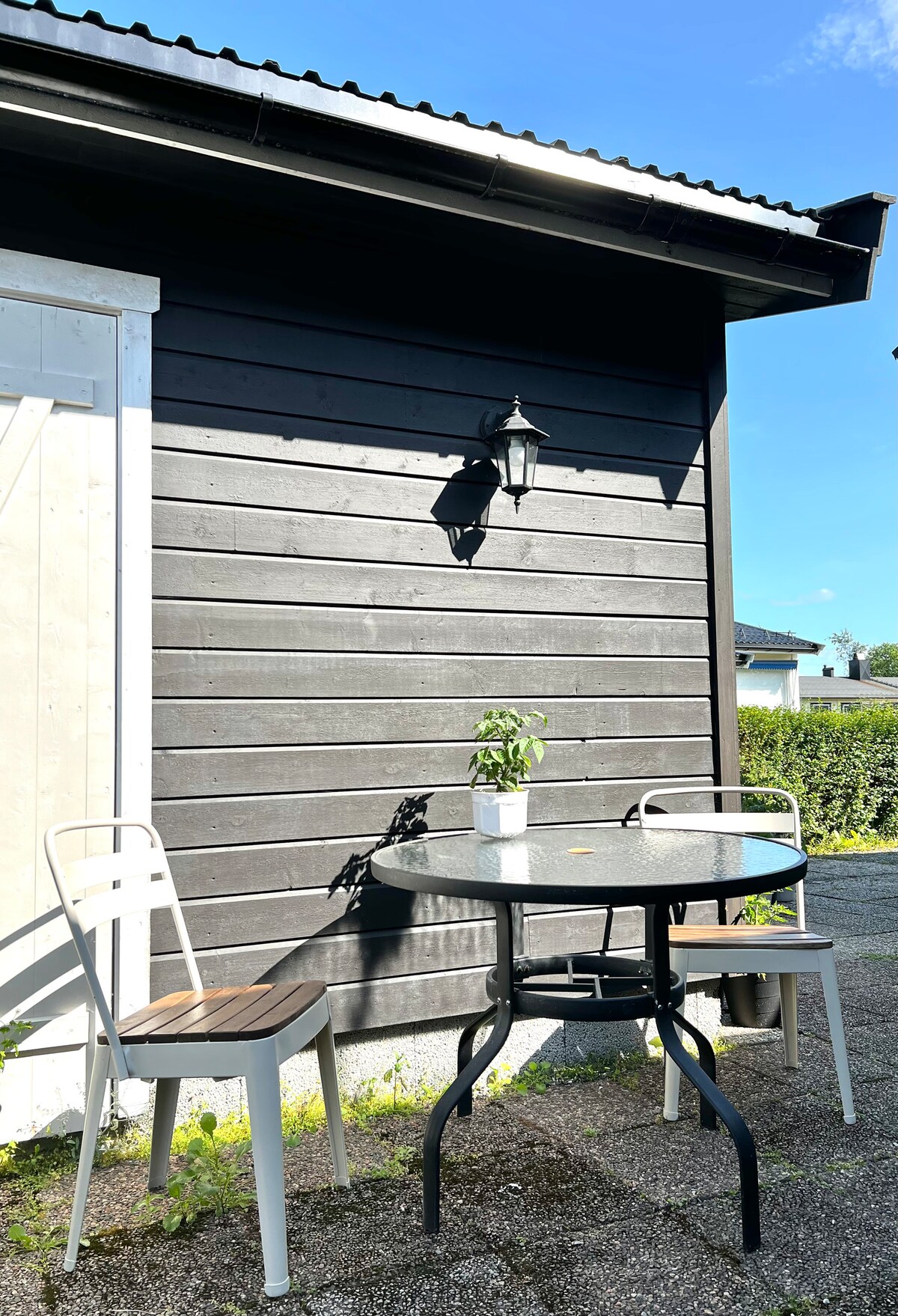 A small outdoor seating area features a round table surrounded by two chairs. Sunlight casts gentle shadows on the surface, highlighting a potted plant at the center. The backdrop consists of a dark wooden wall and a lantern, suggesting a quaint and relaxing space.