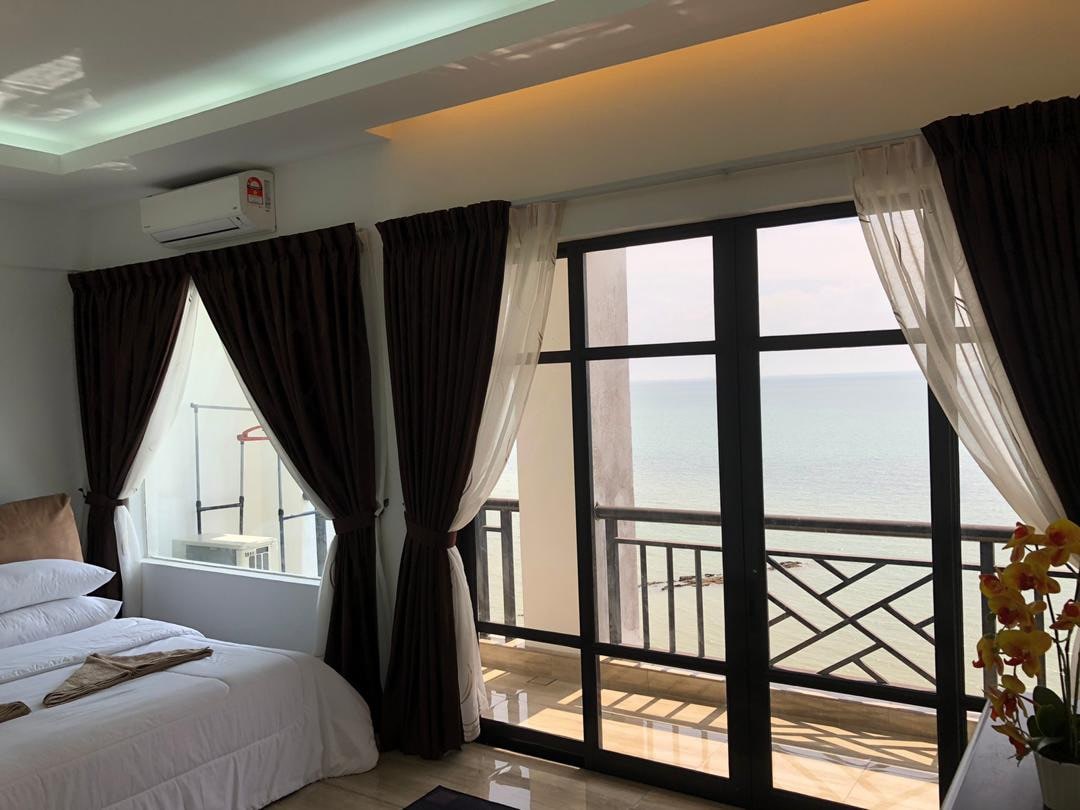 A serene bedroom features a spacious bed with crisp linens. Large glass doors provide a view of the ocean, complemented by sheer and dark curtains. Natural light fills the room, enhancing the overall peaceful ambiance.