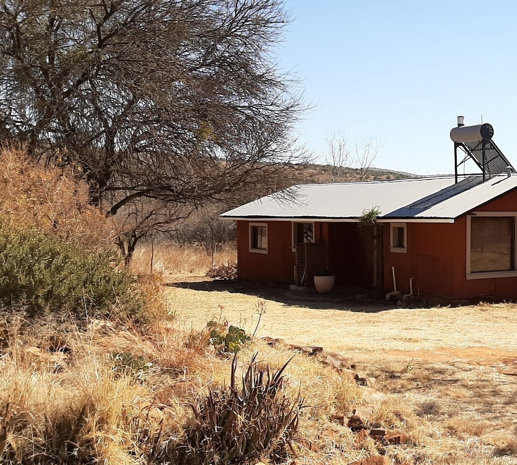 A rustic red exterior of the accommodation is visible, surrounded by dry grass and sparse trees. A water tank is mounted on the roof, and large windows provide natural light to the interior. The landscape features gentle hills in the background.