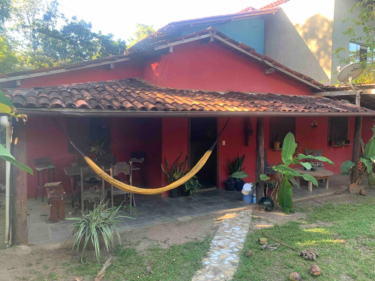 A vibrant red house is framed by lush greenery, featuring a tiled roof and a shaded porch. A yellow hammock swings gently, inviting relaxation. Potted plants adorn the entrance, while a stone pathway leads to the garden area, suggesting a peaceful outdoor space.