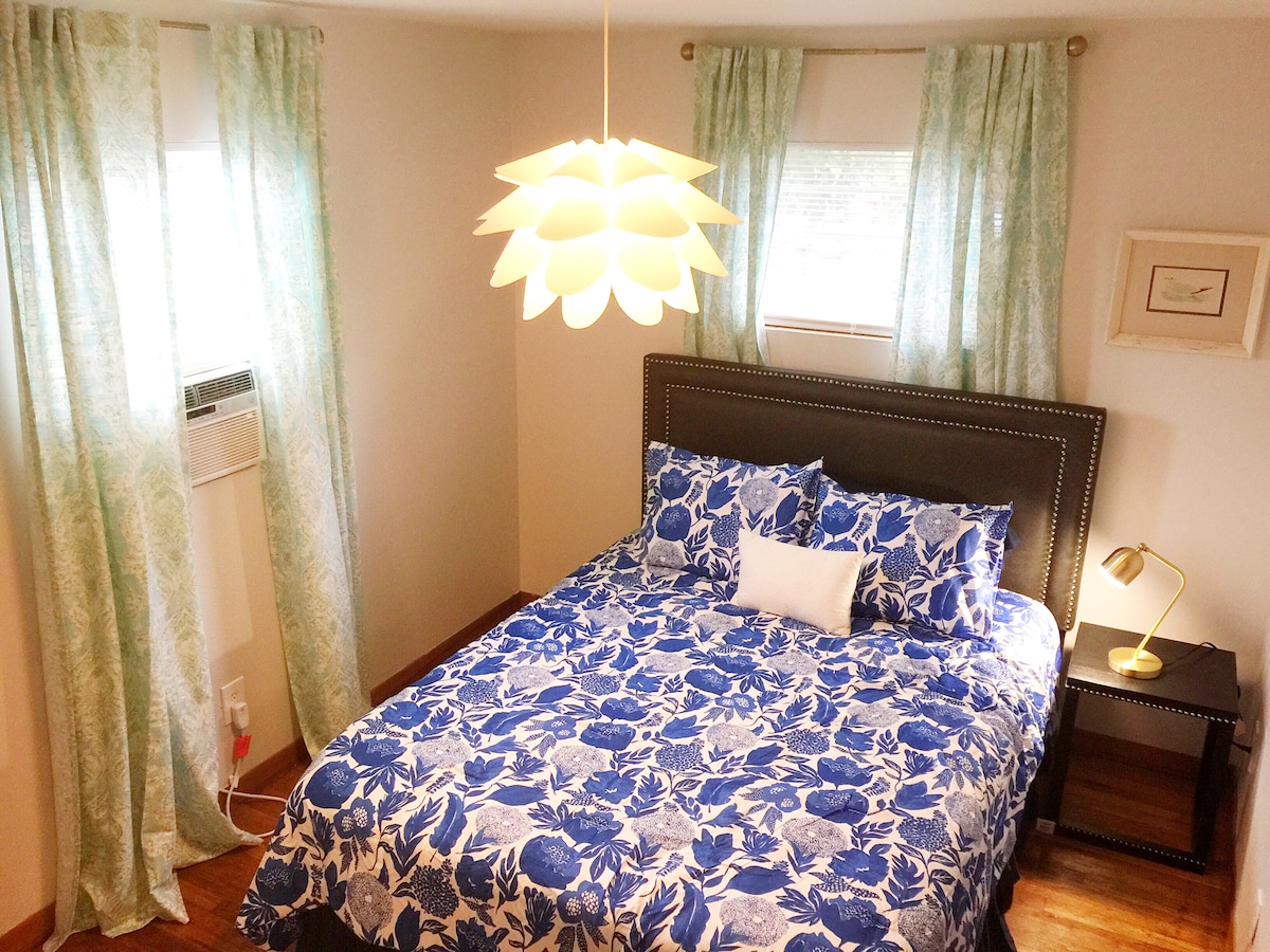 A spacious bedroom is presented with floral-patterned bedding and a decorative pillow. Sheer curtains allow soft light to enter from two windows. A modern pendant light hangs above, and a side table with a lamp is positioned next to the bed, enhancing functionality.