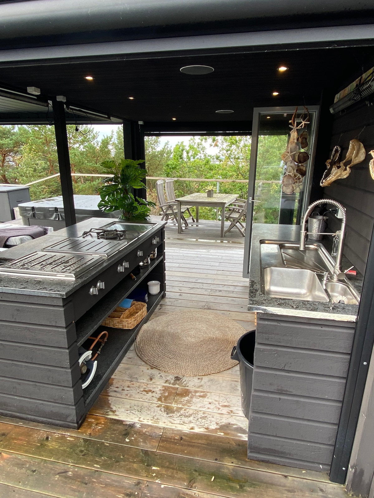 An outdoor kitchen area is presented, featuring a built-in grill and a stainless steel sink. Dark cabinetry complements the granite countertop, while a woven rug adds texture. A dining table and chairs are visible through the glass doors, surrounded by lush greenery.