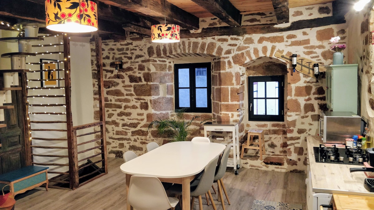 The interior space features exposed stone walls complemented by wooden beams. A dining table surrounded by light-colored chairs is positioned in the center. Two windows provide natural light, and a staircase is visible on the left, leading to the upper area of the home.