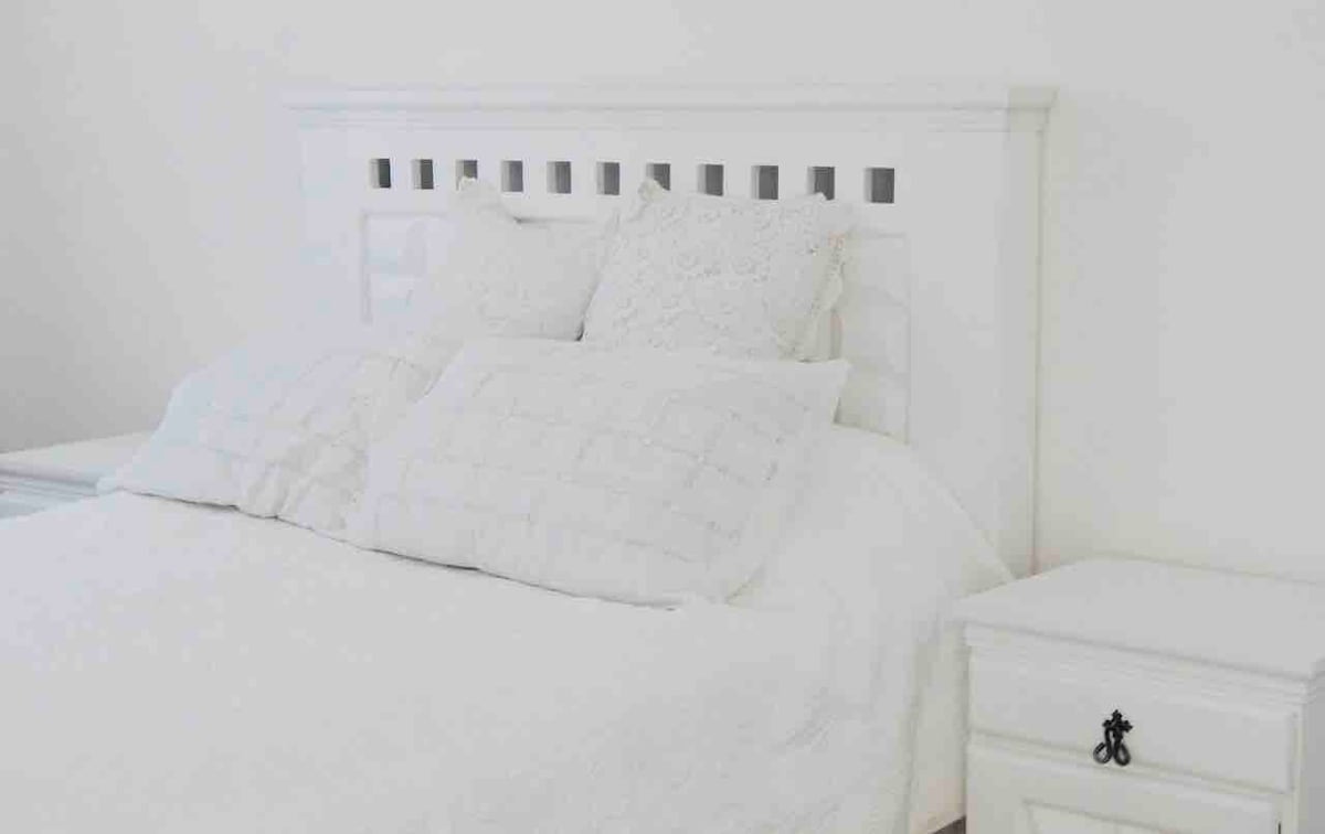A clean, minimalist bedroom showcases a white headboard with decorative detailing. The spacious bed is dressed in soft white linens and multiple textured pillows. A nightstand with a simple design is positioned alongside the bed, contributing to a serene atmosphere.