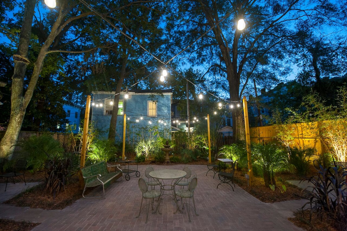 An inviting courtyard is illuminated by softly glowing string lights at dusk. Lush greenery surrounds a central brick-paved patio, featuring a round table and chairs, inviting outdoor gatherings. Tall trees provide shade and a natural backdrop, enhancing the tranquil atmosphere.
