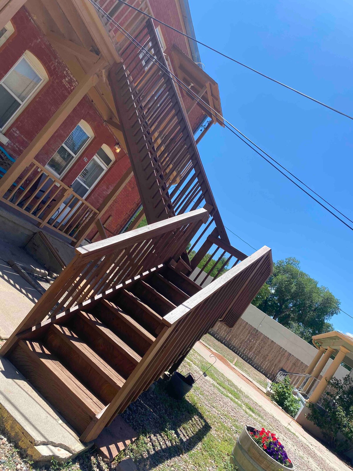 An outdoor staircase leads to the second floor of a brick building. The wooden steps feature a sturdy railing, while blue skies and bright sunlight create a cheerful ambiance. Surrounding greenery is subtly visible in the background.
