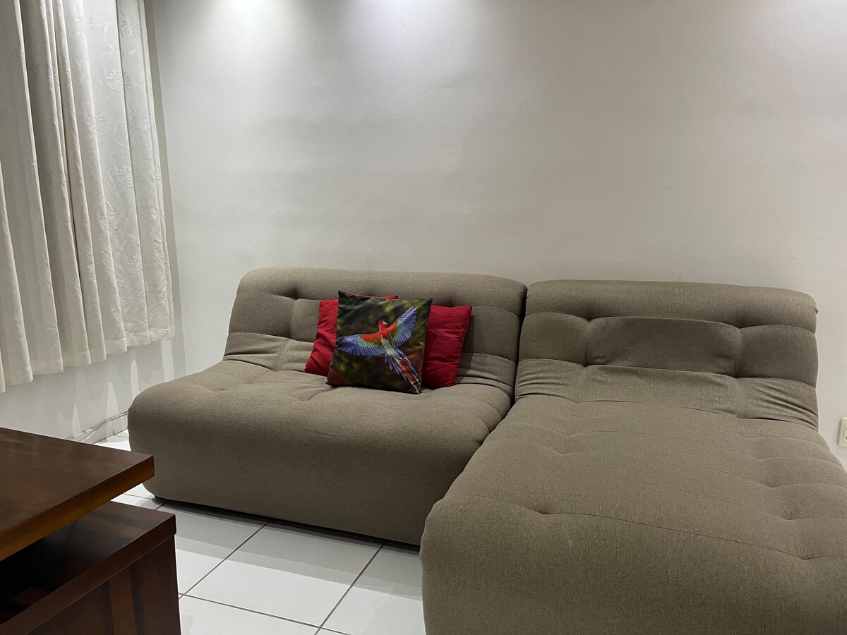 A comfortable sectional sofa is situated in a well-lit living area, featuring a decorative cushion with a colorful design. The simple, neutral walls are complemented by soft drapes, allowing natural light to enhance the space.