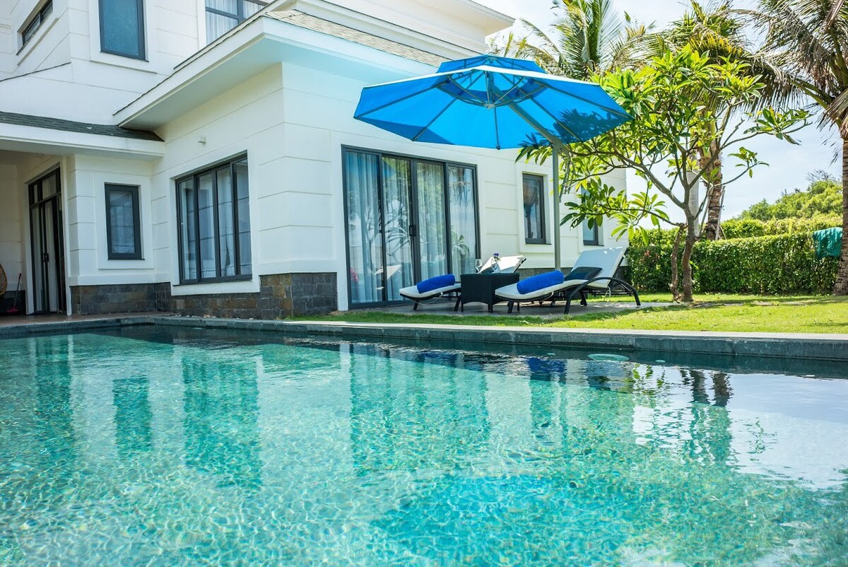 A spacious private pool is bordered by a well-manicured garden, with comfortable lounge chairs positioned nearby. A large blue umbrella provides shade, complementing the clean lines of the modern villa in the background, which features expansive glass doors and a welcoming exterior.