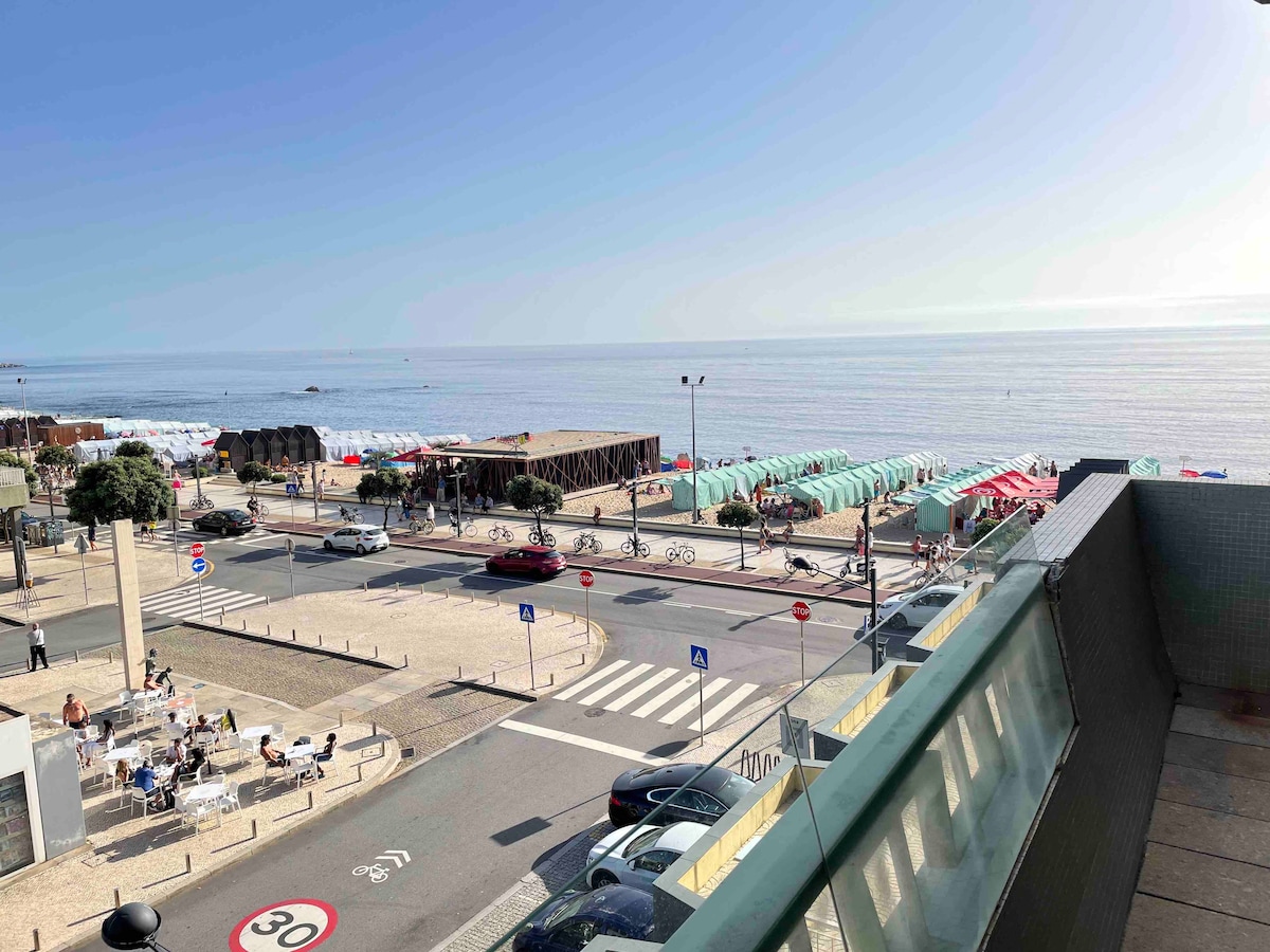 A sea view is captured from a balcony, showcasing a stretch of ocean meeting the horizon. Beach umbrellas and lounge areas are visible along the coastline. A road runs in front with cyclists, cars, and diners seated at outdoor tables, all set against a clear blue sky.