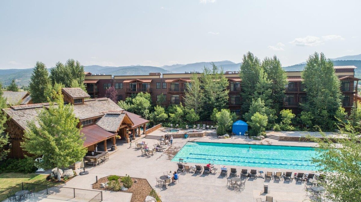 A spacious outdoor area features a large swimming pool surrounded by sun loungers and tables. Lush greenery and trees frame the pool, while a building of rustic design stands in the background, providing a scenic backdrop of the mountains.
