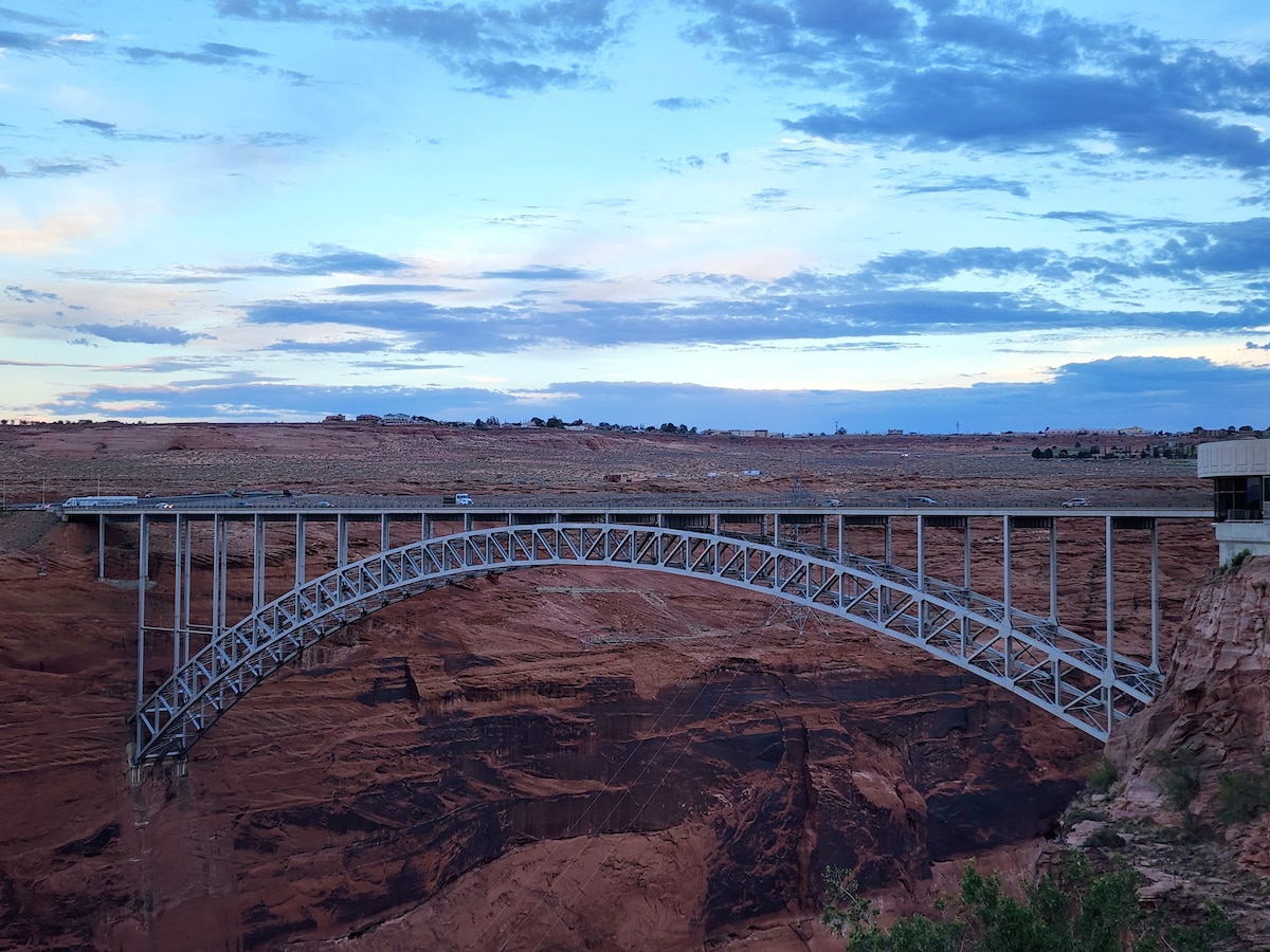 A large steel arch bridge spans a deep canyon, with rocky red cliffs rising prominently on either side. The sky is painted with soft hues of blue and pink, suggesting the end of the day. A distant road is visible atop the bridge.