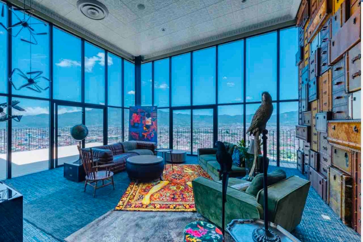 A sunlit living area features large windows offering panoramic views of the mountains and cityscape. Various seating options, including a green couch and a wooden chair, are placed on a colorful area rug. Artistic decor, including a vibrant wall piece, enhances the space.