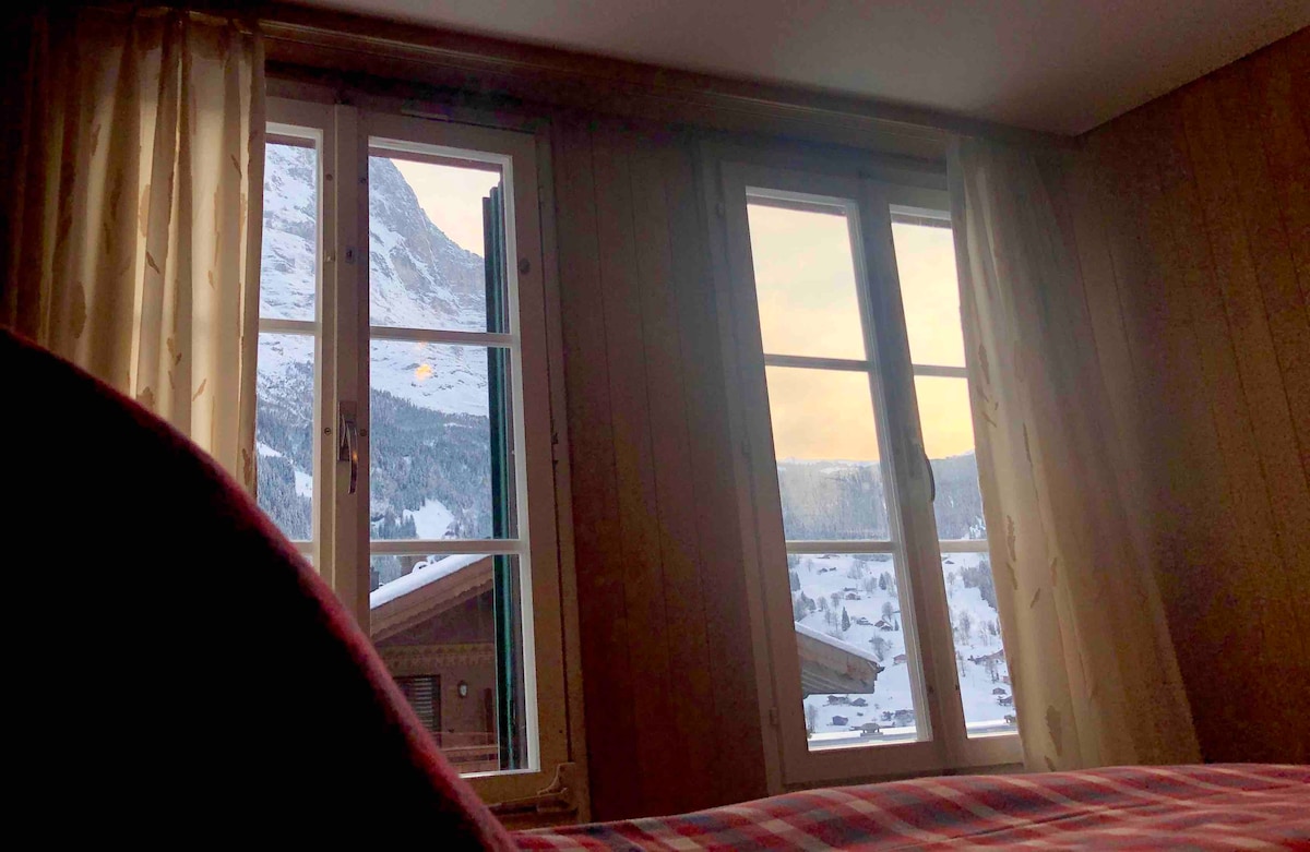 Two large windows frame a view of snow-covered mountains, allowing natural light to fill the room. Soft curtains drape beside the windows, complementing the warm wooden paneling. A cozy sleeping area is visible, with a blanket casually draped over the spacious bed.