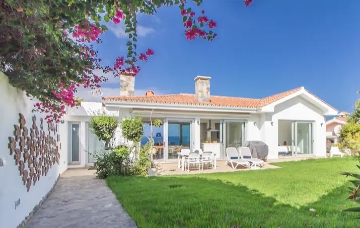 The exterior of the villa features a well-maintained garden with lush green grass and flowering plants. Large windows and glass doors provide ample natural light, showcasing views of the sea. A patio area includes sun loungers and a dining table, inviting outdoor relaxation.