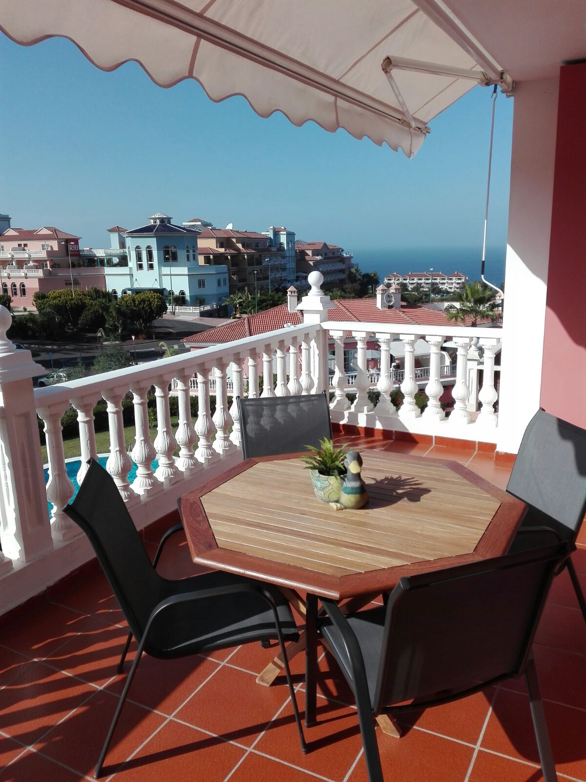 A spacious terrace is shown, featuring a wooden octagonal table accompanied by two black chairs. A small plant adorns the table, and the ocean can be seen in the background, along with distant buildings under a clear blue sky.