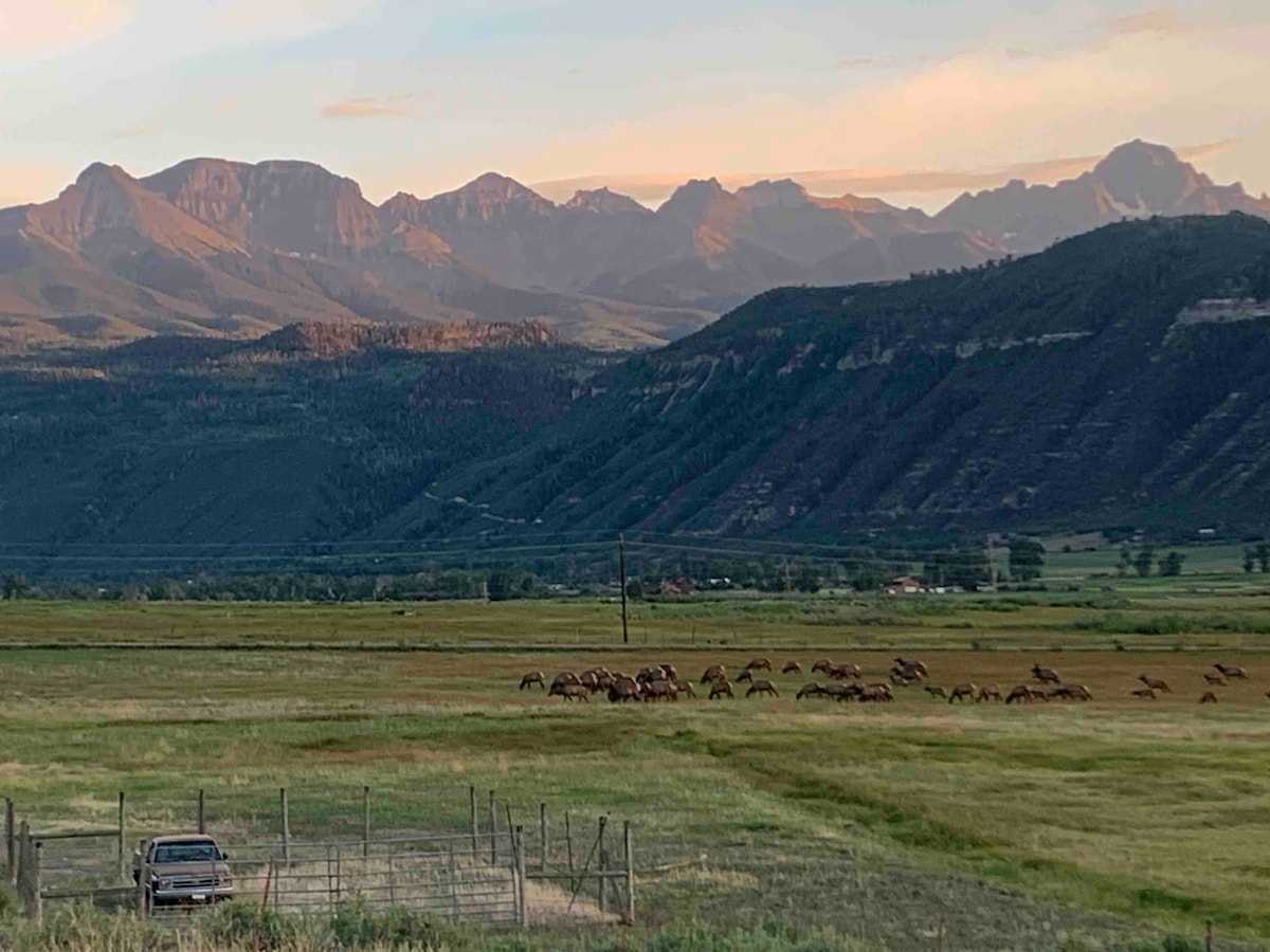 A scenic view of rolling fields under a sunset sky, with mountains in the background. A herd of deer is seen grazing across the grassy area, complementing the natural landscape. Fencing lines the foreground, providing context to the expansive ranch setting.