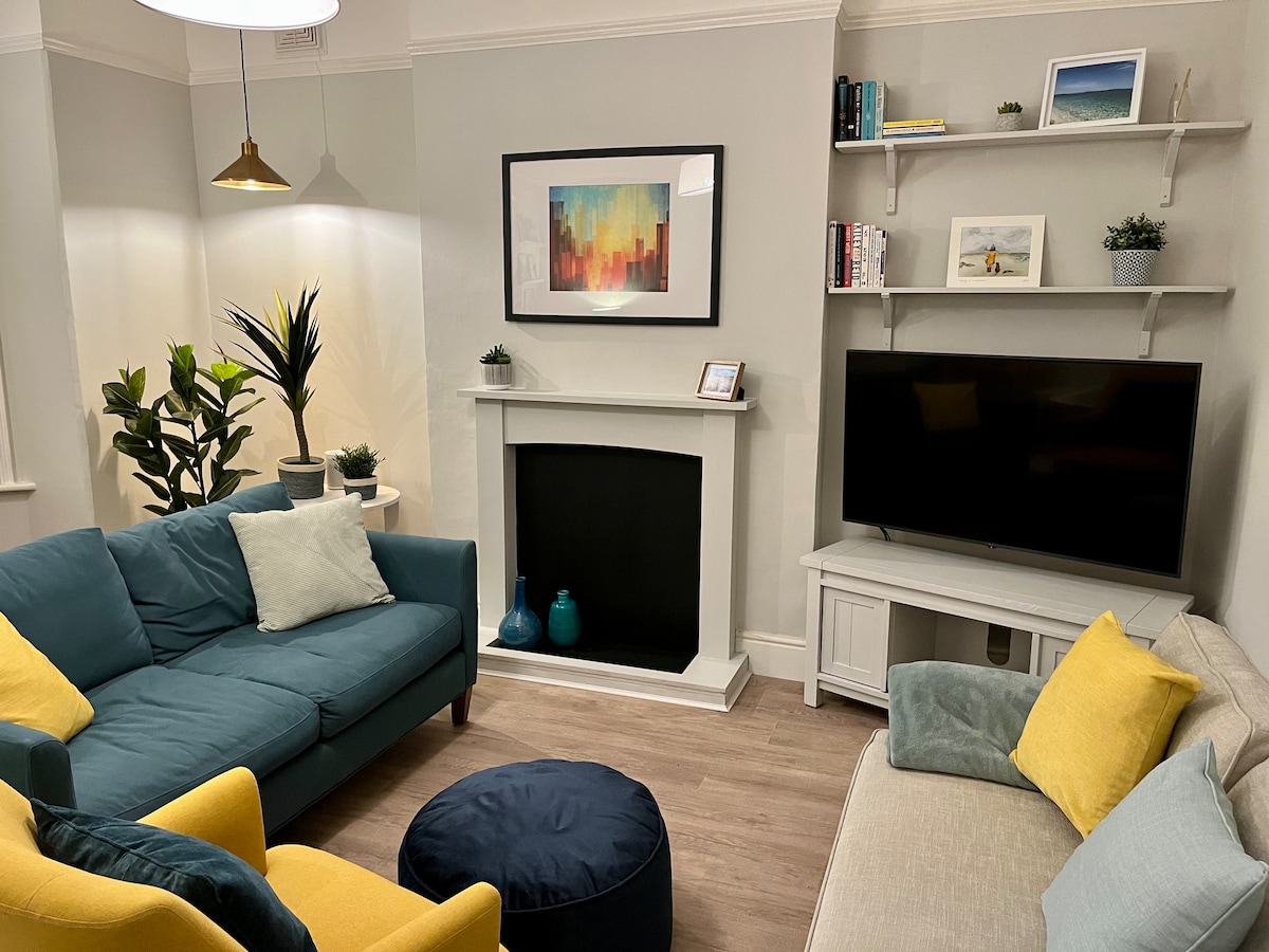 A comfortable living room is depicted with a mix of seating featuring a blue sofa and two yellow chairs. A small round ottoman sits on the floor, and a television is placed beside a white entertainment unit. Decorative plants and artwork enhance the space.