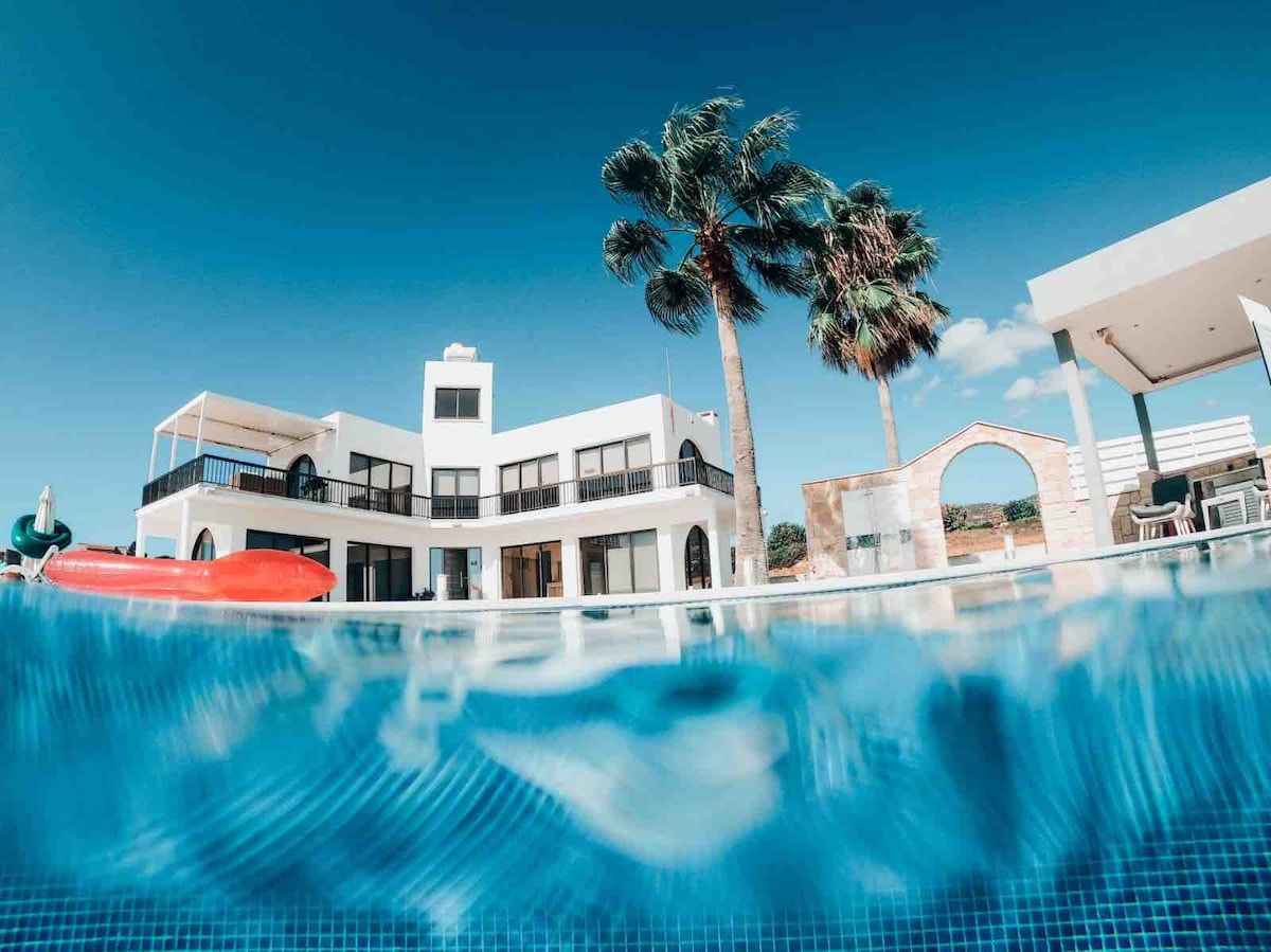 A view of the villa is captured from the perspective of the newly constructed infinity pool. The modern white building features multiple balconies, surrounded by tall palm trees, with clear blue skies above. Red kayaks rest on the pool's edge, enhancing the inviting outdoor space.
