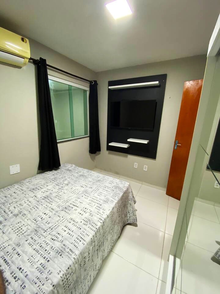 The bedroom features a spacious bed covered with a patterned blanket. A 32-inch smart TV is mounted on the wall, and natural light filters through a window, which is framed by dark curtains. A mirror-fronted wardrobe provides additional storage.