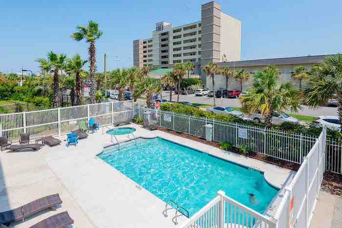 A pool area is surrounded by a fence, featuring a clear blue swimming pool and a nearby hot tub. Luxurious lounge chairs are positioned around the pool, complemented by palm trees that provide a tropical feel. A multi-story building is visible in the background.