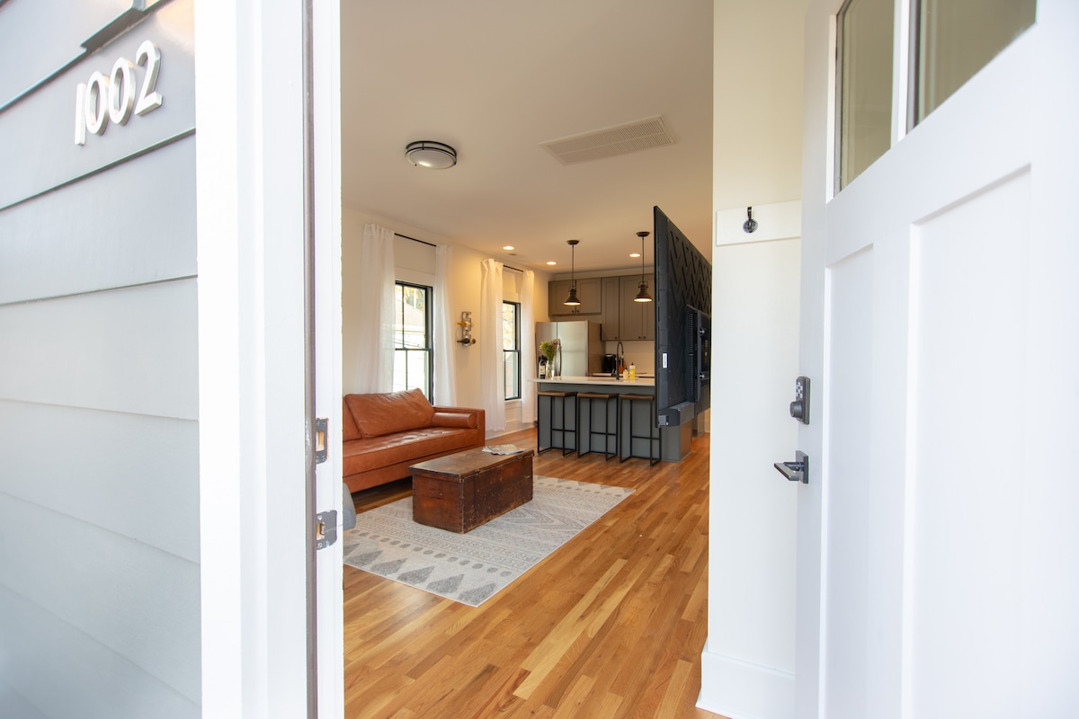 A welcoming entryway leads to an open living space. A leather sofa is positioned beside a wooden coffee table, with a patterned rug beneath. A kitchen island with seating is visible in the background, complementing the hardwood flooring throughout.