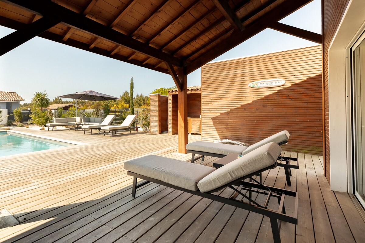 Two loungers are positioned under a wooden pergola, allowing for relaxation beside the heated salt pool. The surrounding area features a landscaped garden with palm trees, enhancing the serene outdoor setting. Sunlight casts gentle shadows on the deck, creating a calm atmosphere.