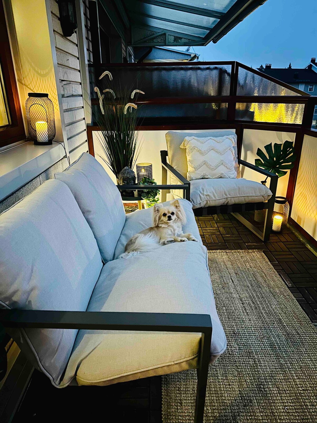 An inviting balcony area features a comfortable, light-colored sofa and matching chair. Soft lighting from lanterns creates a cozy ambiance. A plant and decorative elements enhance the space, while a small dog is comfortably seated on the sofa, adding a touch of warmth to the scene.