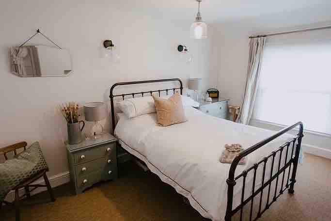A cozy double bedroom features a metal bed frame with crisp white bedding. A decorative pillow is placed at the head of the bed, while a wooden nightstand holds a small vase of flowers. Natural light filters through a large window, complemented by neutral-toned curtains.