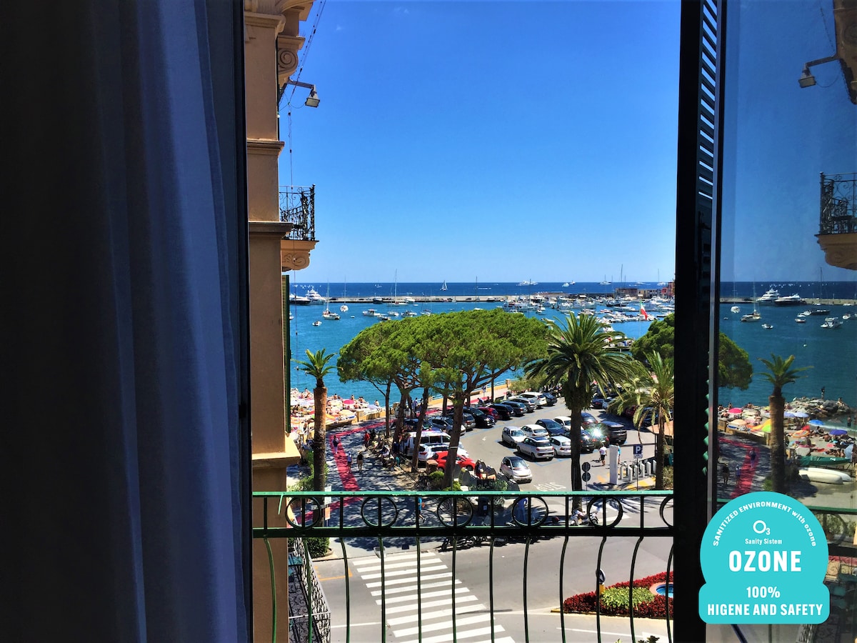 A view through an open window reveals a vibrant waterfront scene, featuring palm trees and boats in the water. Sunlit terraces with colorful umbrellas are visible, indicating a lively beach atmosphere. The deep blue sea stretches out toward the horizon under clear skies.