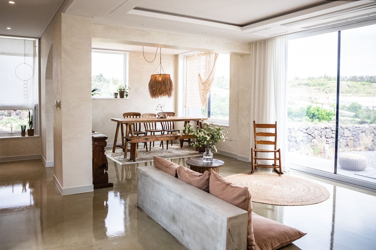 A light-filled living area is showcased with large windows offering views of the surrounding landscape. The space features a wooden dining table, a comfortable rocking chair, and a seating area with soft cushions on a concrete bench. Natural textures are emphasized with a woven pendant light and rug.