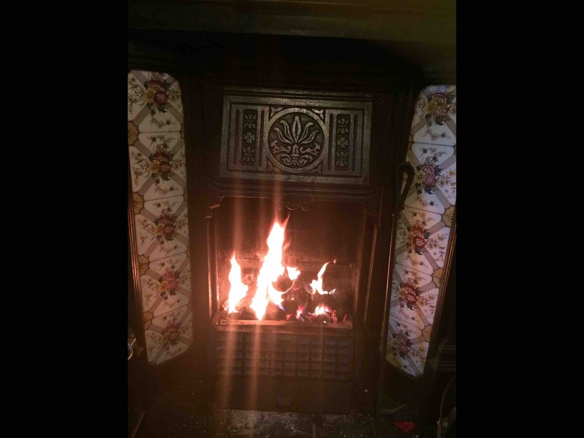 A traditional fireplace is set against intricately patterned tiled surrounds, showcasing flickering flames that provide warmth. The ornate detailing of the fireplace adds character, while the glow of the fire creates an inviting ambiance in the room.