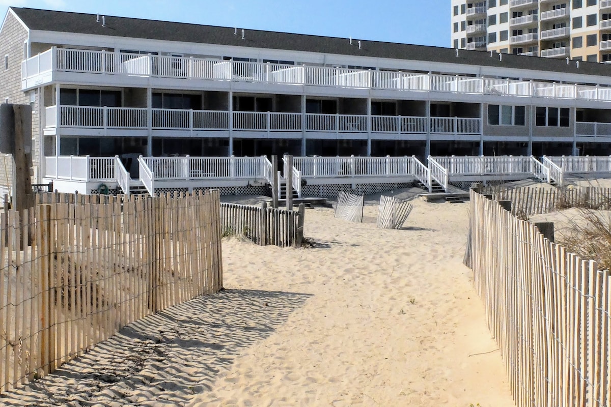 The beachfront property is depicted, featuring a two-story structure with multiple balconies. White railings line the expansive decks, overlooking the sandy beach. Sunlight brightens the scene, highlighting the natural surroundings and the wooden fencing that frames the pathway to the beach.