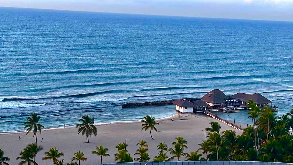 A serene view of the Caribbean Sea can be seen, with gentle waves lapping at the shore. Palm trees line the sandy beach, and a wooden structure is visible at the ocean's edge, creating a tranquil coastal scene.