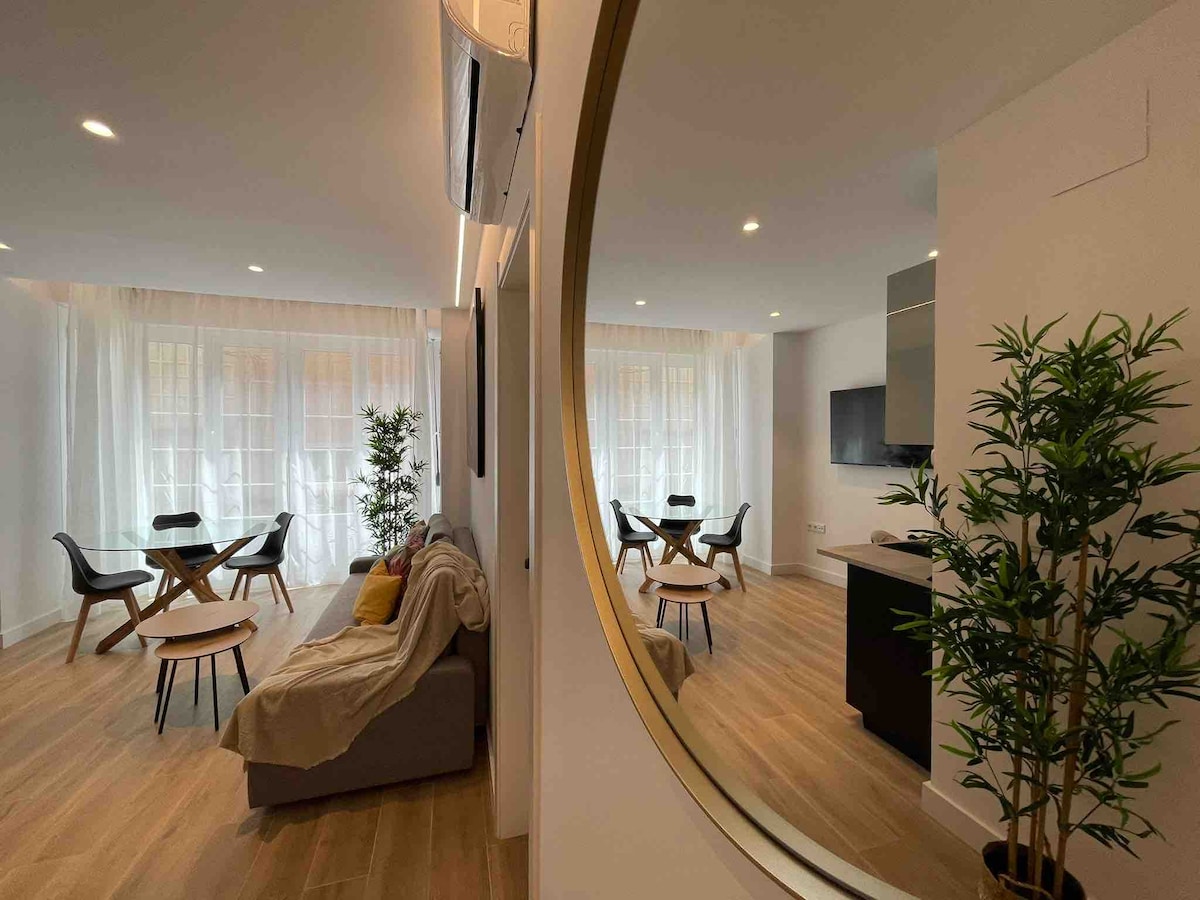 A spacious living area is showcased, with large windows allowing natural light to fill the room. A comfortable sofa is featured alongside a dining table with chairs. A wall-mounted television is visible, with decorative plants adding a touch of greenery to the space.