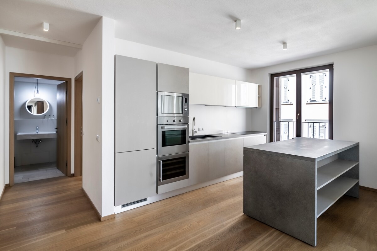 A modern kitchen space is visible, featuring sleek cabinetry and integrated appliances, including an oven and a refrigerator. The neutral color palette complements the natural wood flooring. Light floods in through two large windows, providing views of the exterior, while a small table with open shelving is positioned centrally.