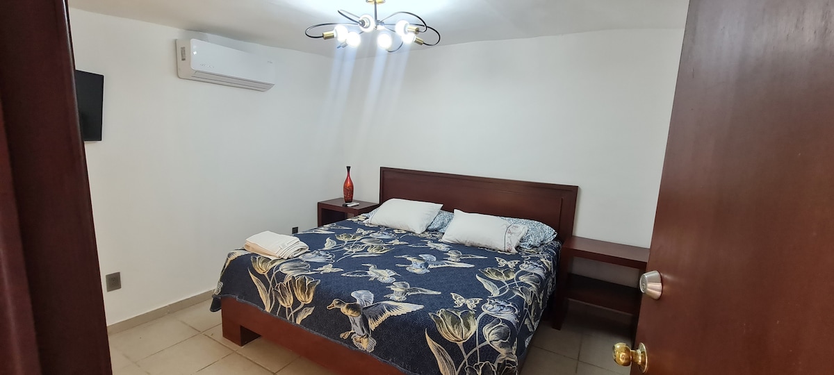 A king-sized bed is centered in the room, adorned with a floral-patterned blanket. Two pillows are placed at the head of the bed. A bedside table with a lamp is positioned beside the bed, and a wall-mounted television is visible. Natural light is gently let in through nearby windows.