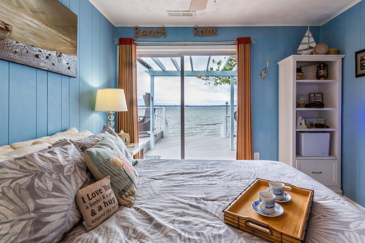 A cozy bedroom features a spacious bed adorned with soft linens and decorative pillows. Natural light enters through sliding glass doors that open to a deck overlooking the water. A bedside table holds a lamp, with a tray presenting two cups beside a light snack.