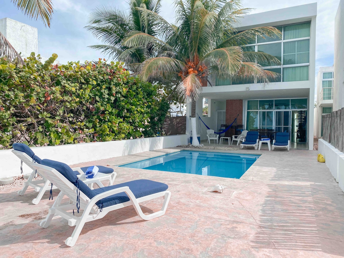 A private outdoor area features a swimming pool surrounded by a spacious patio. Blue lounge chairs are positioned near the water, complemented by tropical palm trees and vibrant greenery. The backdrop includes large glass doors, providing a view into the interior living space.