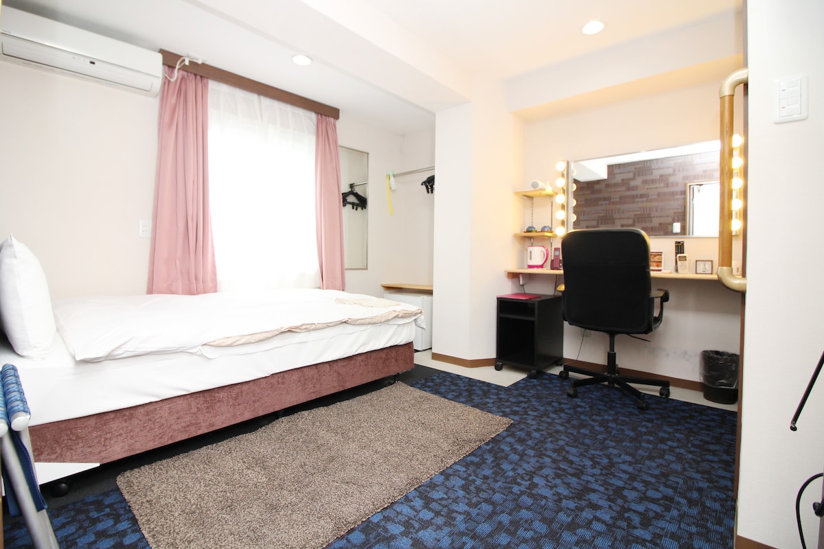 The private room features a semi-double bed with a light blanket on a textured rug. A vanity with lights and a chair is present, offering a space for personal grooming. Large windows dressed in pink curtains allow natural light, illuminating the room's neutral decor.