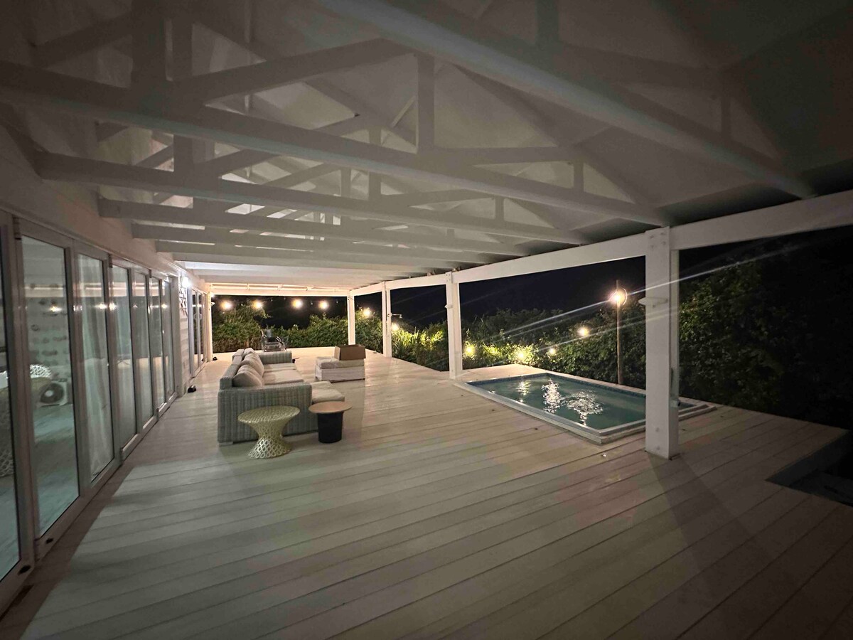A spacious, open patio is illuminated by string lights, featuring comfortable seating and a small table. A private pool is visible nearby, with lush greenery surrounding the area. Large sliding glass doors offer access to the interior space.