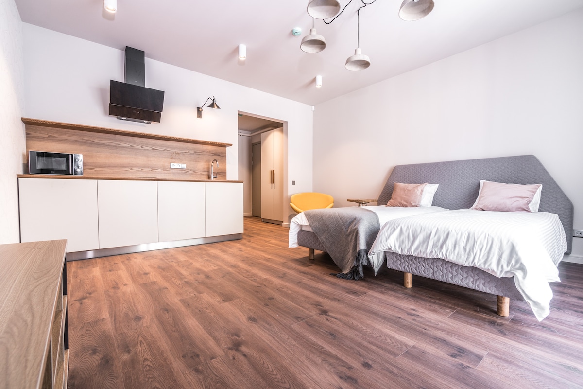 A modern studio space features a stylish kitchen area with built-in cabinetry and a microwave. Two spacious beds are framed by soft textiles, accented by two pastel pillows. Warm wooden flooring enhances the inviting environment, while ceiling lights provide a bright atmosphere.