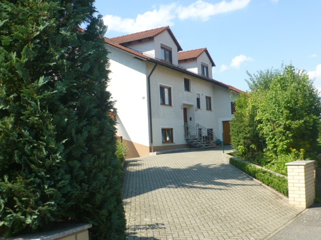 The exterior of the house showcases a two-story structure with a light-colored facade and a sloped roof. Greenery surrounds the property, with hedges and trees framing the entrance. A paved parking area is visible in the foreground, leading to the entry steps.