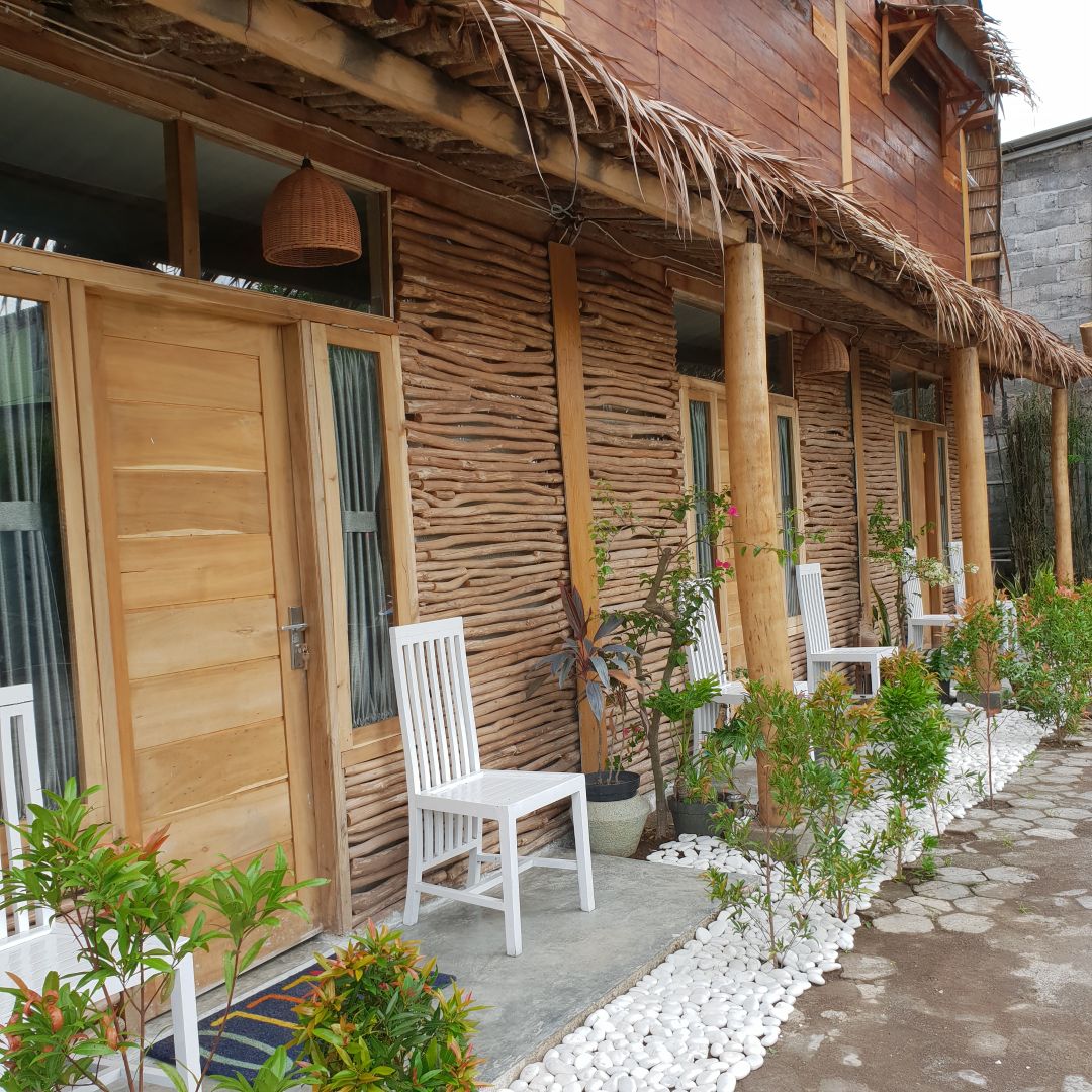 A welcoming entrance showcases a rustic wooden facade with natural elements. Two white chairs are positioned on a stone path, flanked by small potted plants and white gravel. The overall design reflects a harmonious blend of traditional and natural aesthetics.